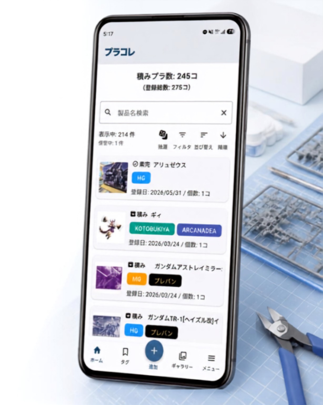 プラモデル制作机の上のスマートフォンに、プラコレのホーム画面(積みプラ数とプラモデル一覧)が表示されている